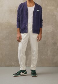 Man wearing white pants, dark green and black sneakers, white tank top, and navy blue buttoned jacket with green Nike logo stands indoors.