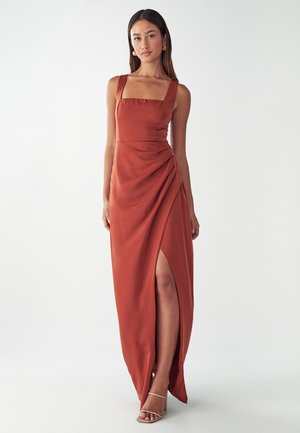 Rust-colored maxi dress with a square neckline, draped design, side slit, and smooth texture. Styled with strappy sandals.