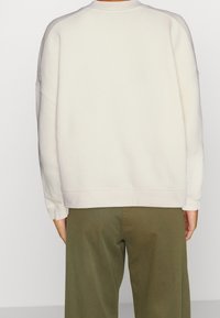 Persona vistiendo un sudadera blanca de manga larga y pantalones verdes olivo, mostrada desde atrás contra un fondo simple.