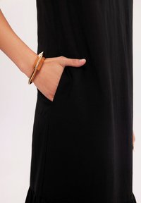 Czarna sukienka z boczną kieszenią, charakteryzująca się gładką teksturą. Na nadgarstku widnieje złocona bransoletka w kształcie geometrycznym.