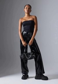 Top preto sem alças, calças pretas brilhantes de cintura alta em verniz, sapatos de plataforma pretos e robustos, e uma pequena mala preta com detalhes em corrente prateada.