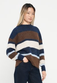 Pull rayé dans des nuances de marron, crème et bleu. Fabriqué en tissu doux et texturé avec un col montant et une coupe ample. Les manches longues sont accentuées de couleurs.