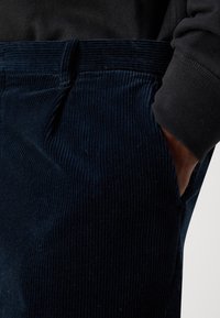 Main dans la poche d'un pantalon en velours côtelé bleu foncé porté avec une chemise noire à manches longues, montrant la texture du tissu et les détails de couture.