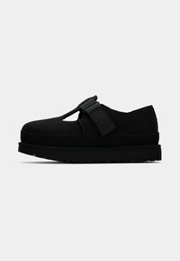 Chaussure plate noire avec une semelle épaisse, découpe ouverte sur le dessus et large bride réglable au niveau du cou-de-pied sur un fond uni.