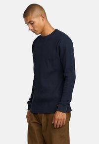 Navy-langarm-Waffle-Strickshirt mit gerippter Textur, rundem Halsausschnitt und kontrastierenden Bündchen. Kombiniert mit sandfarbenen Hosen, getragen von einem Modell.