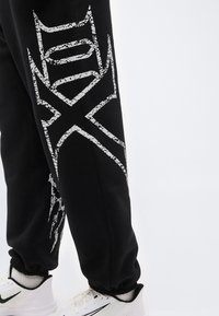 Μαύρο παντελόνι jogging με μεγάλο, ραγισμένο λευκό γραφικό σχέδιο στο πόδι, φορεμένο με λευκά αθλητικά παπούτσια Nike και λευκές κάλτσες πάνω σε απλό φόντο.