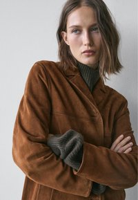 Bruine suède jas met een gladde textuur, voorzien van een kraag, knopen aan de voorkant en grijze gebreide mouwen die tevoorschijn komen.