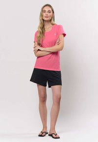 Ružové tričko s krátkym rukávom a okrúhlym výstrihom s malým logom, v kombinácii s čiernymi šortkami a žabkami, na neutrálnom pozadí.