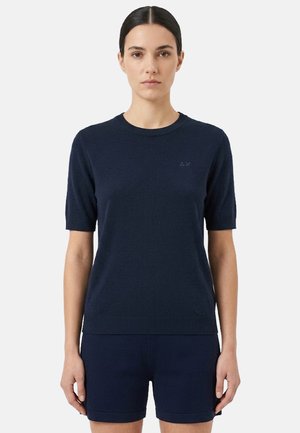 Donna con capelli scuri, indossa un top in maglia a maniche corte blu navy e pantaloncini abbinati blu navy, in piedi davanti a uno sfondo chiaro e semplice.