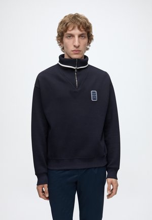 Junger Mann mit kurzen lockigen Haaren, der einen marineblauen Pullover mit hohem Reißverschlusskragen und kleinem Logo auf der Brust trägt, vor neutralem Hintergrund stehend.
