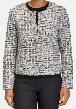 Mujer con chaqueta de tejido de tweed en blanco y negro con cuello redondo y sin botones visibles, combinada con pantalones negros ajustados.