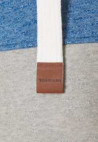 Sweatshirt mit einem oberen Teil aus blauem, strukturiertem Strick und einem grauen unteren Abschnitt, ausgestattet mit einer weißen, gewebten Kordel und einem braunen Lederetikett mit Prägung „TOM TAILOR“.