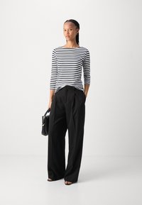 Schwarz-weiß gestreiftes Langarmoberteil kombiniert mit schwarzen weiten Hosen. Das Model hält eine schwarze Handtasche und zeigt einen lässigen, aber eleganten Look.