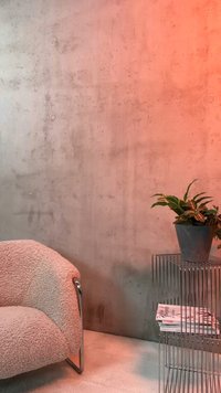 Zakrivljena bež fotelja pokraj žičanog metalnog pomoćnog stolića na kojem stoji posađeno zeleno bilje i složeni časopisi uz betonski zid.