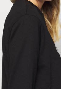 Sweatshirt noir avec un col rond côtelé, couture d'épaule visible et texture lisse. Présente un design imprimé sur le devant.