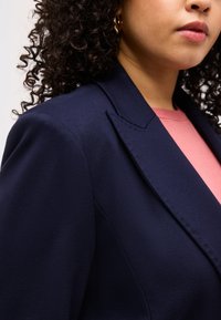 Γυναίκα με σγουρά μαλλιά που φοράει χρυσό κρίκο στα αυτιά, μπλε σκούρο σακάκι και ροζ τοπ, εμφανιζόμενη από τον ώμο έως τα χείλη σε ουδέτερο φόντο.