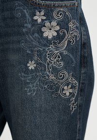 Σκούρο μπλε τζιν με ανοιχτό γκρι κεντήματα λουλουδιών που απεικονίζουν λουλούδια και στροβιλιζόμενα φύλλα κατά μήκος της περιοχής του μηρού.