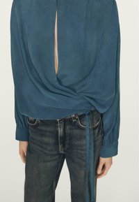 Blågrøn bluse med draperet design, udskæring på ryggen og bundet detalje ved taljen, kombineret med mørke cowboybukser. Glat stoftekstur.