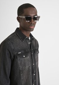Camisa de mezclilla negra con dos bolsillos en el pecho y botones plateados, llevada con gafas de sol rectangulares translúcidas con lentes oscuros.