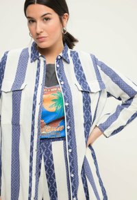 Camicia a righe blu e bianche con motivi, caratterizzata da un design con bottoni frontali e due tasche sul petto, indossata sopra una maglietta grafica.