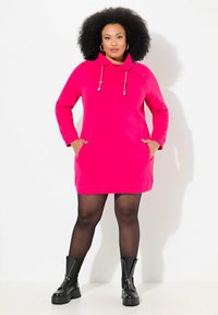 Vestido sudadera rosa con cuello de cisne, detalles de cordón, mangas largas y bolsillos laterales, combinado con botas negras de tobillo y medias.