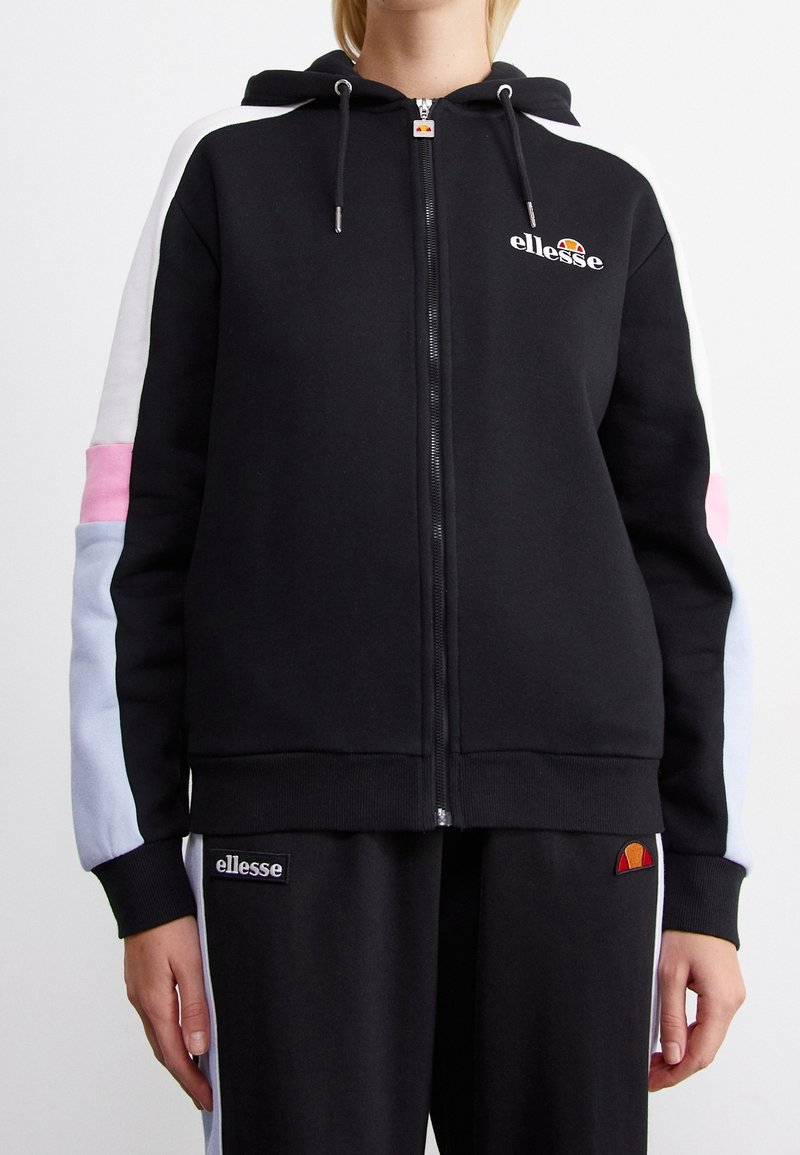 Sweat à capuche noir zippé avec des panneaux de manches blancs, roses et bleu clair, associé à un pantalon noir avec des logos ellesse sur la poitrine et la cuisse.
