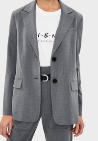 Kvinna som bär en grå skräddarsydd kavaj och matchande byxor med ett svart bälte, i kombination med en vit grafisk T-shirt med texten "Television S."