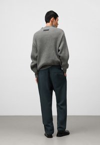 Maglione grigio a coste con etichetta nera sul retro, abbinato a pantaloni a gamba larga verde scuro. Calzature nere completano l'outfit.