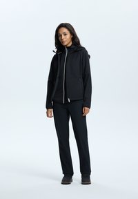 Giacca softshell nera con zip, cappuccio e due tasche; abbinata a pantaloni neri e stivali neri resistenti, con un maglione strutturato sotto.