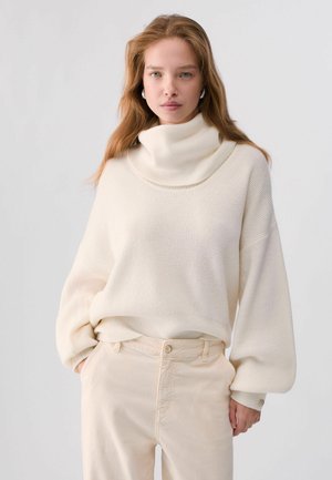 Junge Frau mit langen hellbraunen Haaren, die einen lockeren weißen Rollkragenpullover und beige Hose trägt mit den Händen in den Taschen.