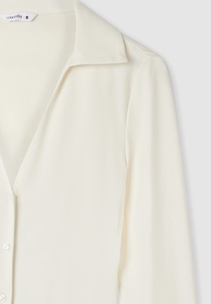 Blusa a maniche lunghe di colore crema con colletto e bottoni sul davanti, mostrata dalla spalla fino a metà torso su uno sfondo chiaro.