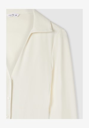 Blusa a maniche lunghe di colore crema con colletto e bottoni sul davanti, mostrata dalla spalla fino a metà torso su uno sfondo chiaro.