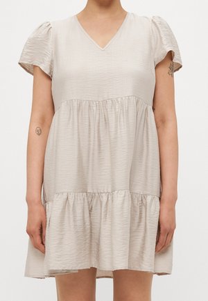 Femme portant une robe courte beige texturée avec des manches courtes bouffantes et une jupe à volants, dévoilant deux petits tatouages sur le bras.