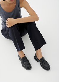 Svarte loafers i skinn med teksturerte detaljer, lav hæl, rund tå og synlige sømmer. Modellen har på seg mørke bukser og en grå topp.