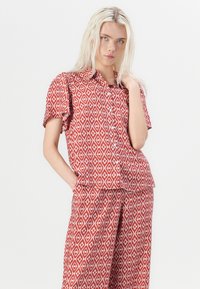 Červený vzorovaný dvojdielny outfit, ktorý obsahuje košeľu s krátkym rukávom a gombíkmi a zodpovedajúce nohavice s diamantovým vzorom. Mäkká textúra látky.