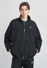 Μαύρο μπουφάν με φερμουάρ και ψηλό γιακά, με αντίθετη γκρι ραφή και μικρό λογότυπο Nike Air στο στήθος. Λεία υφή.