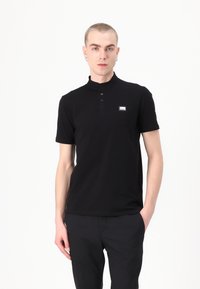 Juodas polo marškinėliai su aukšta apykakle, trumpomis rankovėmis ir mygtukų eilute. Kairėje krūtinės pusėje yra mažas logotipo užrašas. Švelnus, lygus paviršius.