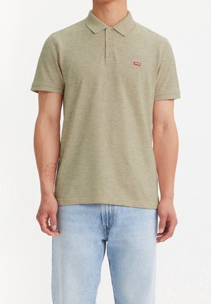 Olive green polo shirt features a classic collar, three-button placket, and a small red logo. Worn with light blue denim jeans.