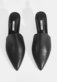 Mules en cuir noir avec un design à bout pointu, texture lisse et sans embellissements. Les semelles sont plates avec le nom de la marque visible à l'intérieur.