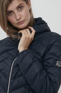 Veste matelassée marine avec design à surpiqûres, capuche et fermeture zippée argentée ; dotée d'un patch logo sur la manche ; matériau lisse et brillant.