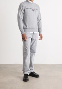 Sudadera gris con logo bordado, combinada con jeans gris claro y zapatos negros sin cordones. Textura suave y diseño ajustado.