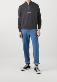 Felpa zip grigia scura con colletto alto e un piccolo logo sul petto, abbinata a jeans azzurri arrotolati e sneaker nere.