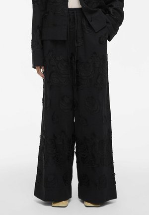 Zwarte wijde broek met textuurlijke bloemenborduursels, gedragen met een bijpassende zwarte jas en beige puntige schoenen.