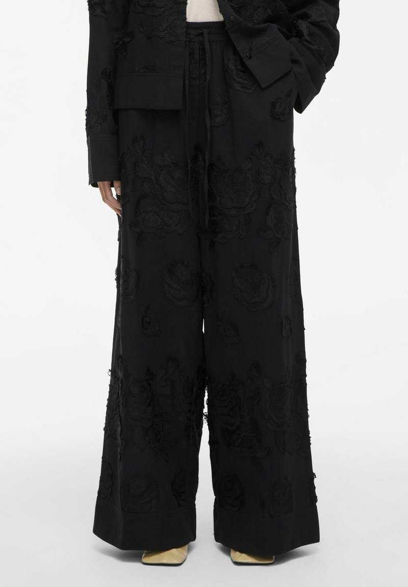 Schwarze weit geschnittene Hose mit strukturiertem floralem Stickmuster, kombiniert mit einer passenden schwarzen Jacke und beigefarbenen spitzen Schuhen.