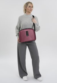 Compact, ribbed, burgundy shoulder bag with a black zipper and branding. Carrying strap shown, made of glossy synthetic material.