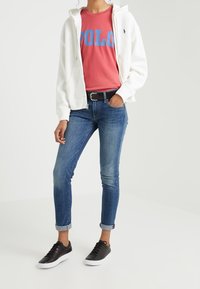 Bílá mikina na zip, růžové tričko s modrým nápisem "POLO", modré úzké džíny a černé tenisky s bílou podrážkou.