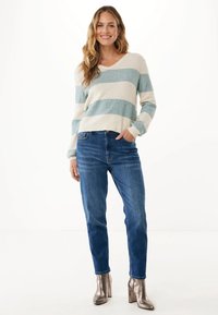 Eine Frau, die lächelt und einen gestreiften Pullover in Weiß und Hellblau, blaue Jeans und metallische, hochhackige Stiefeletten trägt, steht mit einer Hand in der Tasche.