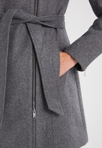 Šedý vlněný kabát s páskem na zavazování, vybavený předním zipem a bočními kapsami. Tkanina má měkkou texturu a střih na míru.