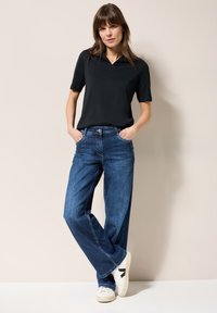 T-shirt polo noir associé à un jean en denim bleu ample et des baskets blanches. Le mannequin se tient avec les mains dans les poches devant un fond neutre.