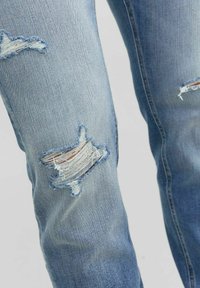 Jeans de denim azul claro con detalles desgastados y rotos en las rodillas, mostrando una textura deshilachada y un ligero desvanecimiento en toda su superficie.
